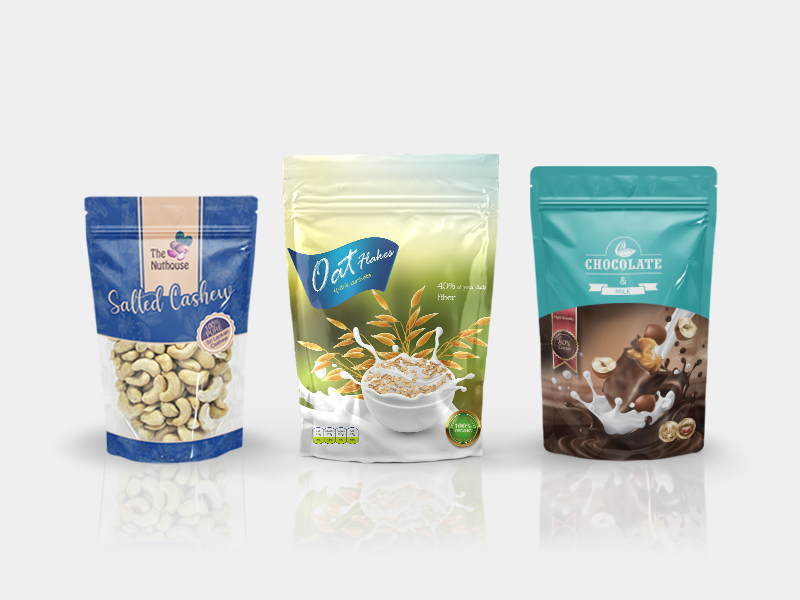 A variety of standing pouches are arranged randomly on a neutral background. A green package labelled-Salted Cashew is transparent, and is filled with whole roasted cashews. Another is labelled-Chocolate and Milk, while a third appears to be labelled-Oats