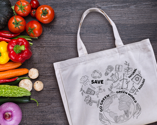 A white canvas tote bag is placed flat on a grey wooden surface, printed with a design in black that says Go Green with illustrations of plants, clouds, and other items. Nearby is a pile of fresh produce.