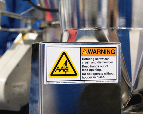 A white colour warning sticker is placed on a metal, printed with a warning message informing workers keep hands out of the feed opening.