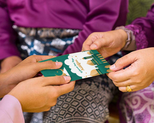 Two hands delicately hold a green-coloured money packet labelled Selamat Hari Raya Aidilfitri, set against a background of individuals dressed in purple clothing.