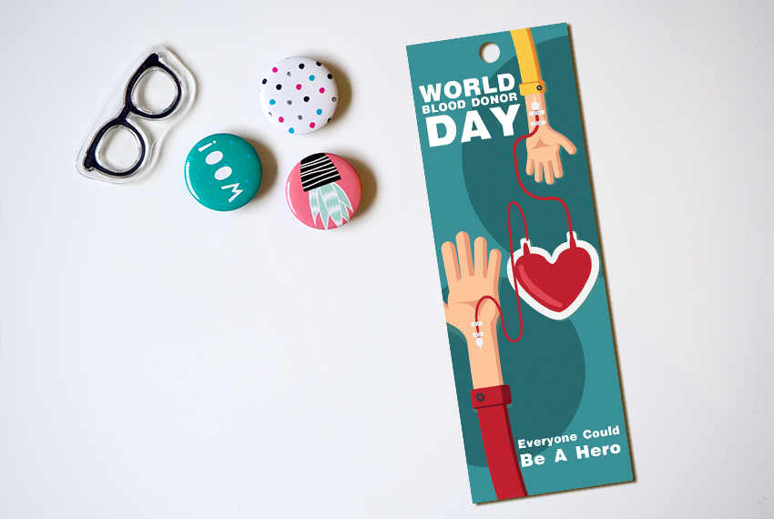 A bookmark with a hole punched out of the top, featuring a stylized cartoon image of a blood drive, placed on a white surface next to three button badges, and a pair of spectacles.
