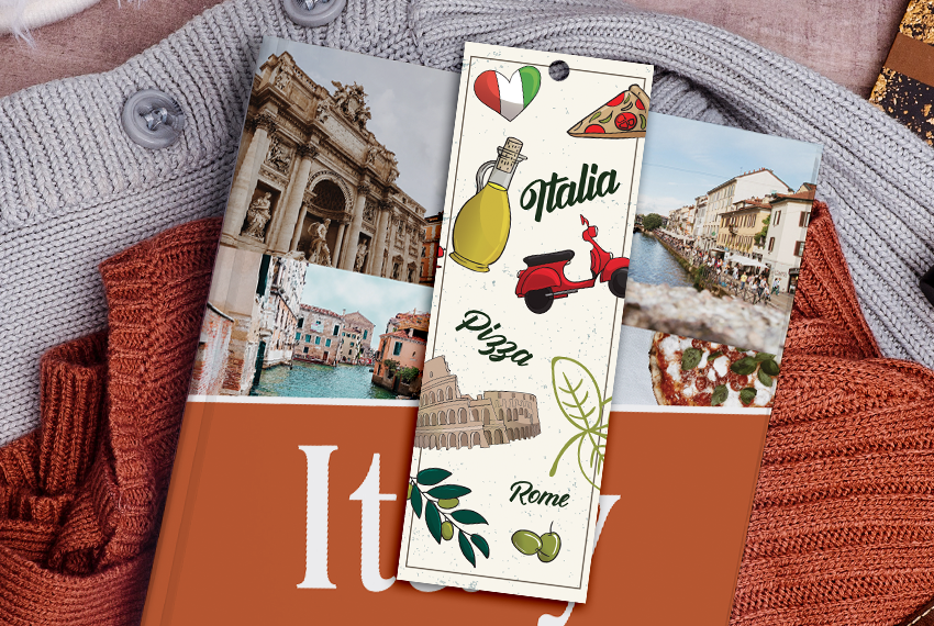 A white, simple bookmark printed with various images of Italian imagery (olives, a Vespa, pizza, etc.), resting on a book about Italy.