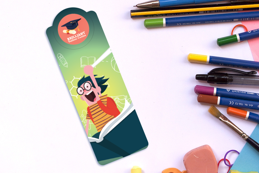A bookmark featuring a cartoon image of a bespectacled individual riding a flying book, placed on a white surface next to an assortment of stationery.