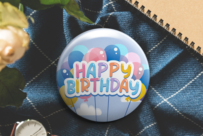 A button badge with an image of a large bunch of balloons set against a blue sky, labelled “Happy Birthday” in pastel colours.