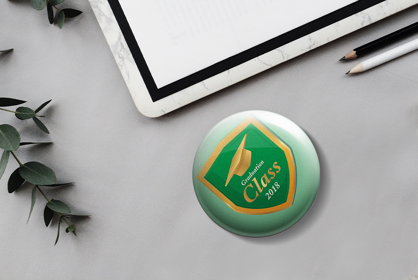 A green button badge decorated with a graduation cap, placed on a grey background next to stationery and leaves.