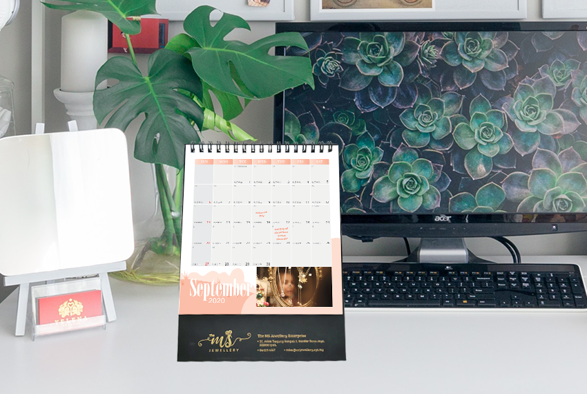 A simple hard stand desk calendar placed on an office desk in front of a desktop computer. A vase holding 3 monstera leaves is sitting in the background next to a small mirror.