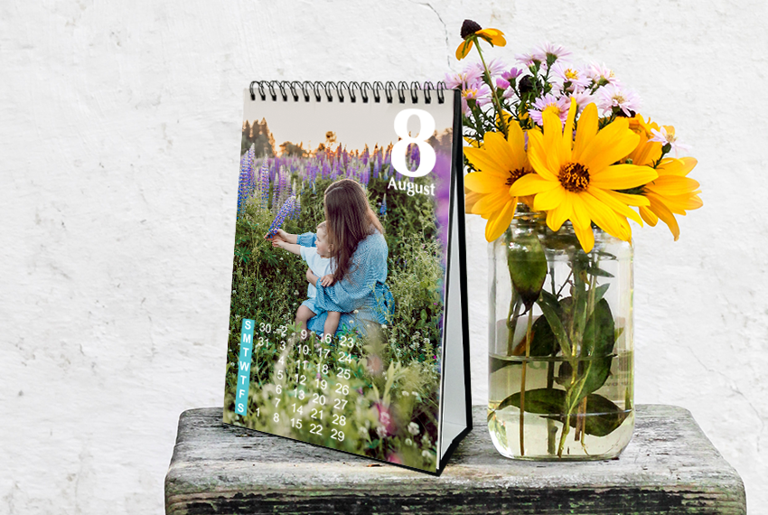 A hard stand desk calendar with the image of a woman and a baby in a field placed next to a jar of flowers, resting on a small table.
