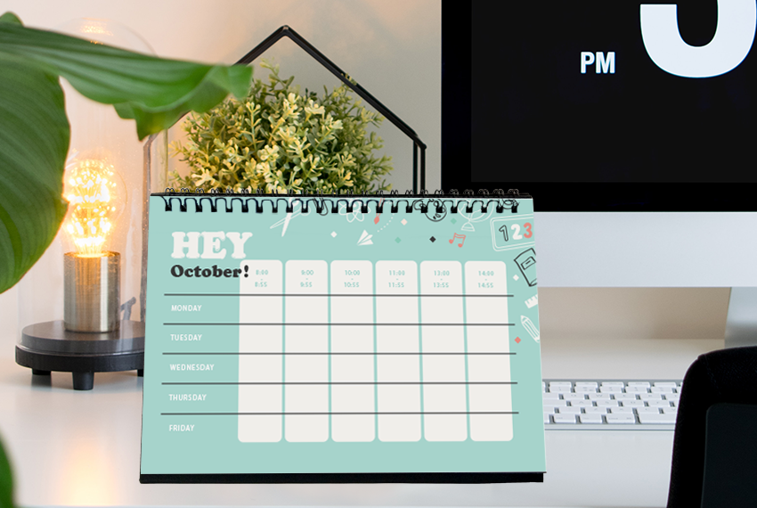 A simple hard stand desk calendar with a light blue background placed on a desk in front of a large Mac computer.