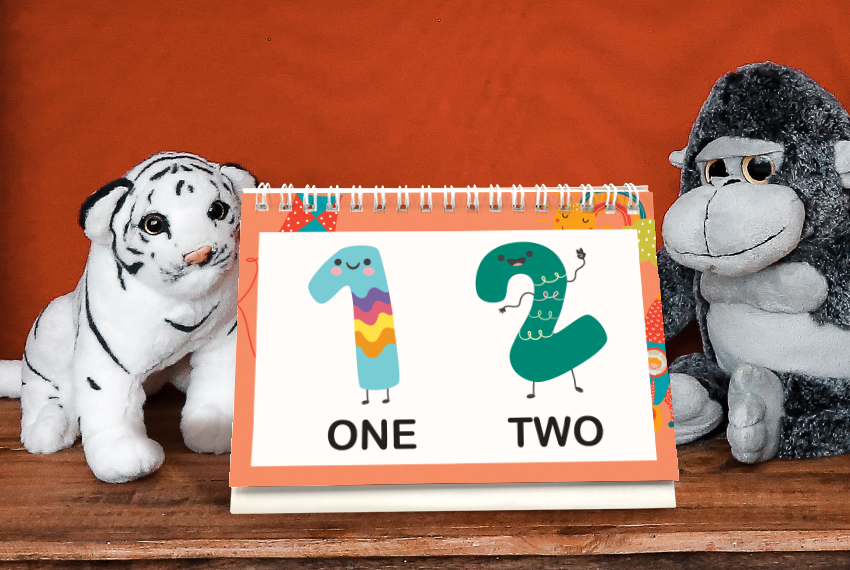 A simple soft stand desk calendar printed with cartoon images of the numbers “1” and “2”, placed in between a plush tiger and a plush gorilla.