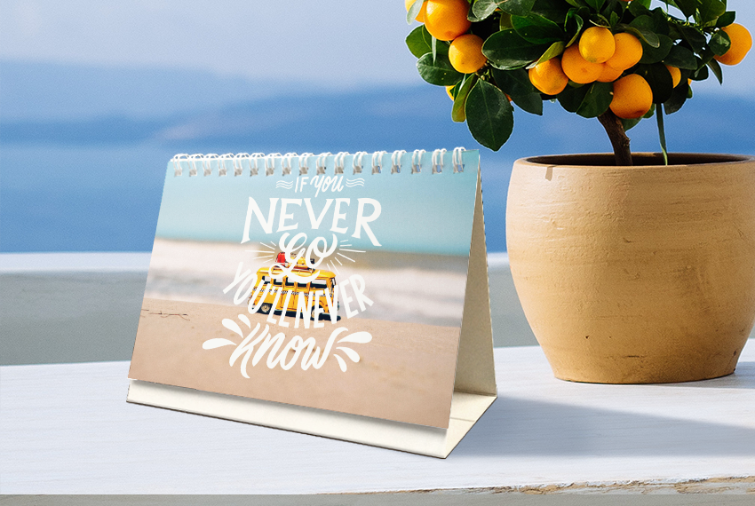 A soft stand desk calendar featuring an image of a small toy on a beach with the quote “If you never go, you’ll never know” printed in white over the image. The calendar is placed next to a citrus plant.