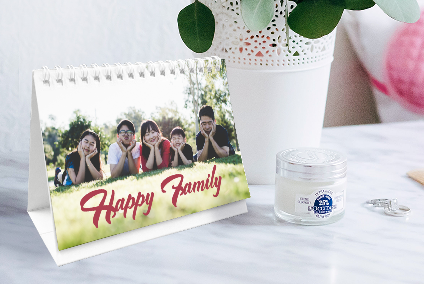 A simple soft stand desk calendar featuring a family photo and labelled “Happy Family” placed on a marble table next to a vase.