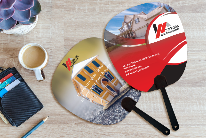 Two hand fans apparently advertising a real estate agency placed on a wooden surface next to a card holder and a cup of coffee.
