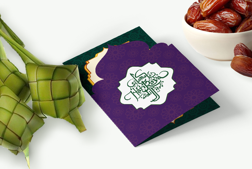 A greeting card for Hari Raya Aidilfitri sits on a white surface next to dates and ketupat.