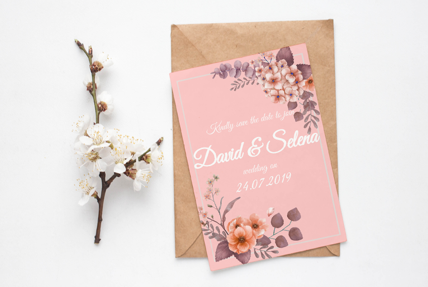 A pink card rests on a brown envelope, apparently designed to be a wedding invitation. There is a cherry blossom branch next to it.