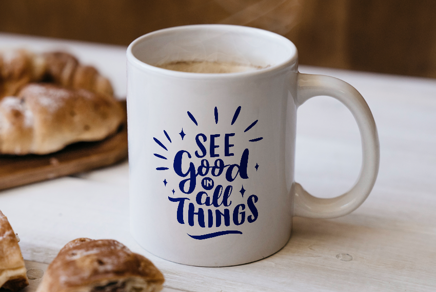 A white mug printed with the text “See Good in All Things” sits on a table next to baked goods.