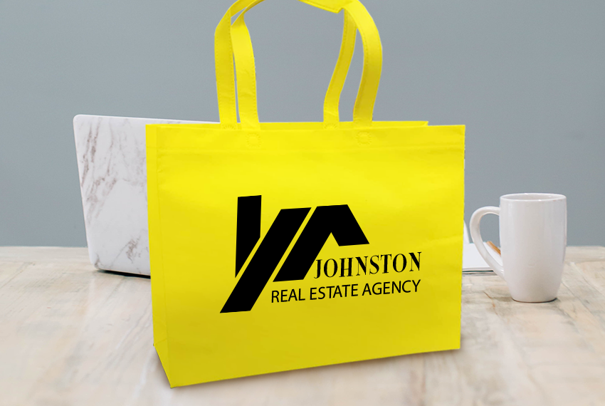 A yellow tote bag printed with “Johnston Real Estate Agency” sits in front of a laptop and mug on a wooden surface.