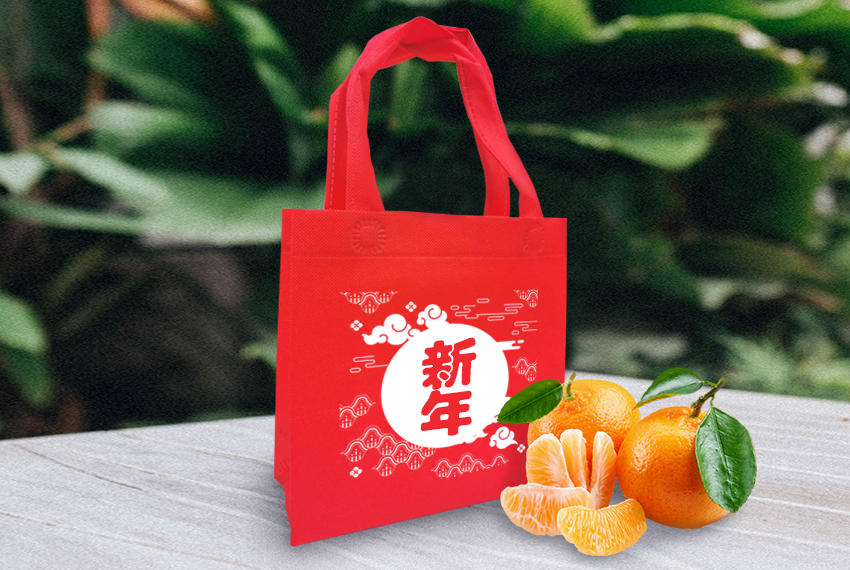 A red tote bag with a Chinese New Year design sits next to a pile of mandarin oranges.