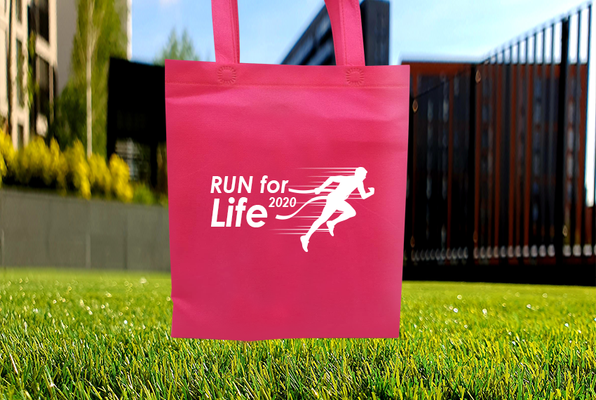 A pink tote bag printed with the words “Run for Life” and the silhouette of a man running past a finish line is held above some grass.