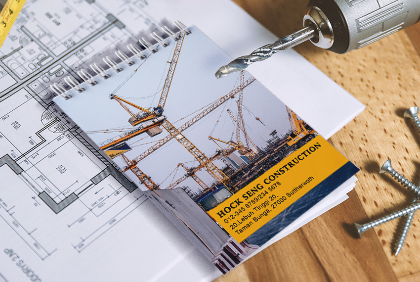 A wirebound notebook sits on top of architecture blueprints, printed with an image of cranes and the words “Hock Seng Construction” at the bottom.