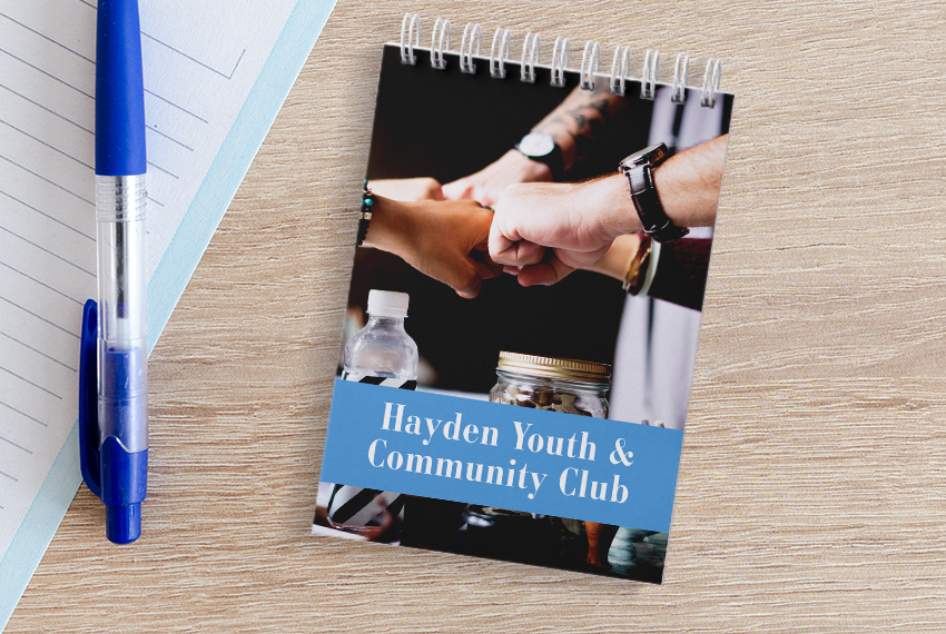 A wirebound notebook printed with “Hayden Youth & Community Club” featuring an image of multiple hands fist-bumping sits on a wooden surface next to another notebook.