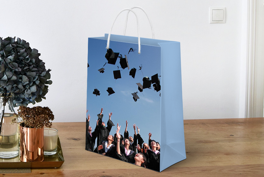 A paper bag featuring the image of a group of graduates throwing their caps into the air sits on a wooden surface next to a small plant.