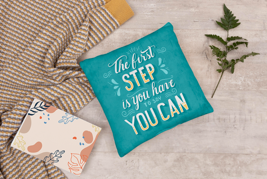 A teal pillow sits on a concrete background. “The first step is you have to say you can” is printed on the pillow. 