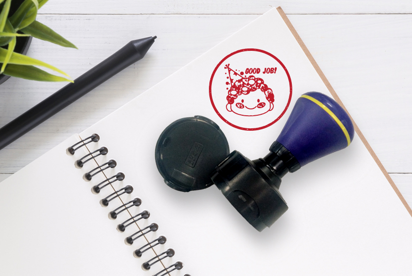 A rubber stamp that says “Good Job” with a smiling face rests on a notebook that has been stamped.
