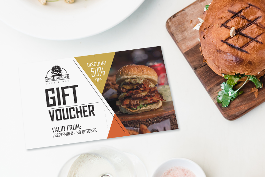 A card printed with “Gift Voucher” and featuring an image of a grilled burger sits on a table next to a burger.