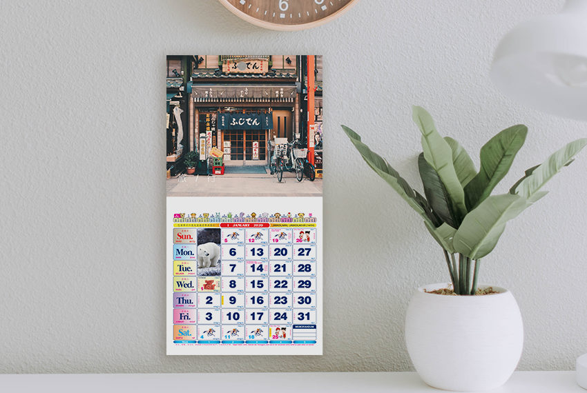 A wall calendar featuring an image of old architecture placed on a white wall next to a potted plant.