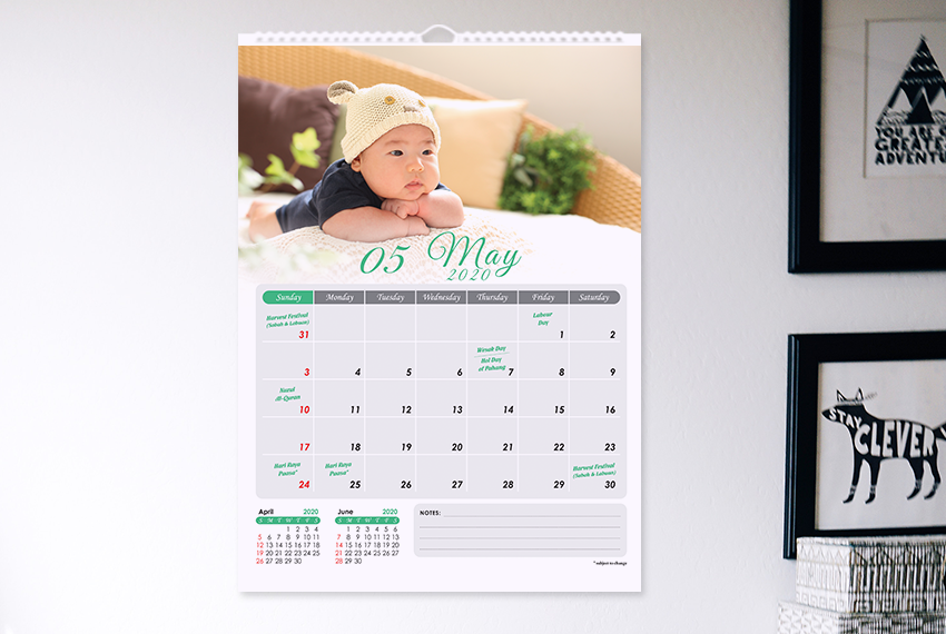 A simple wire-O bound wall calendar featuring the image of a small baby placed on a white wall next to two framed pictures.
